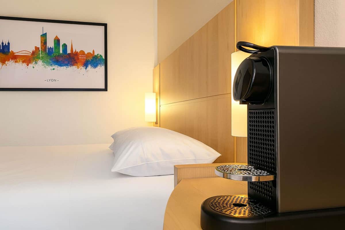 A hotel room with a bed, a pillow, a wall-mounted artwork depicting a city skyline, and a coffee machine on a wooden table.