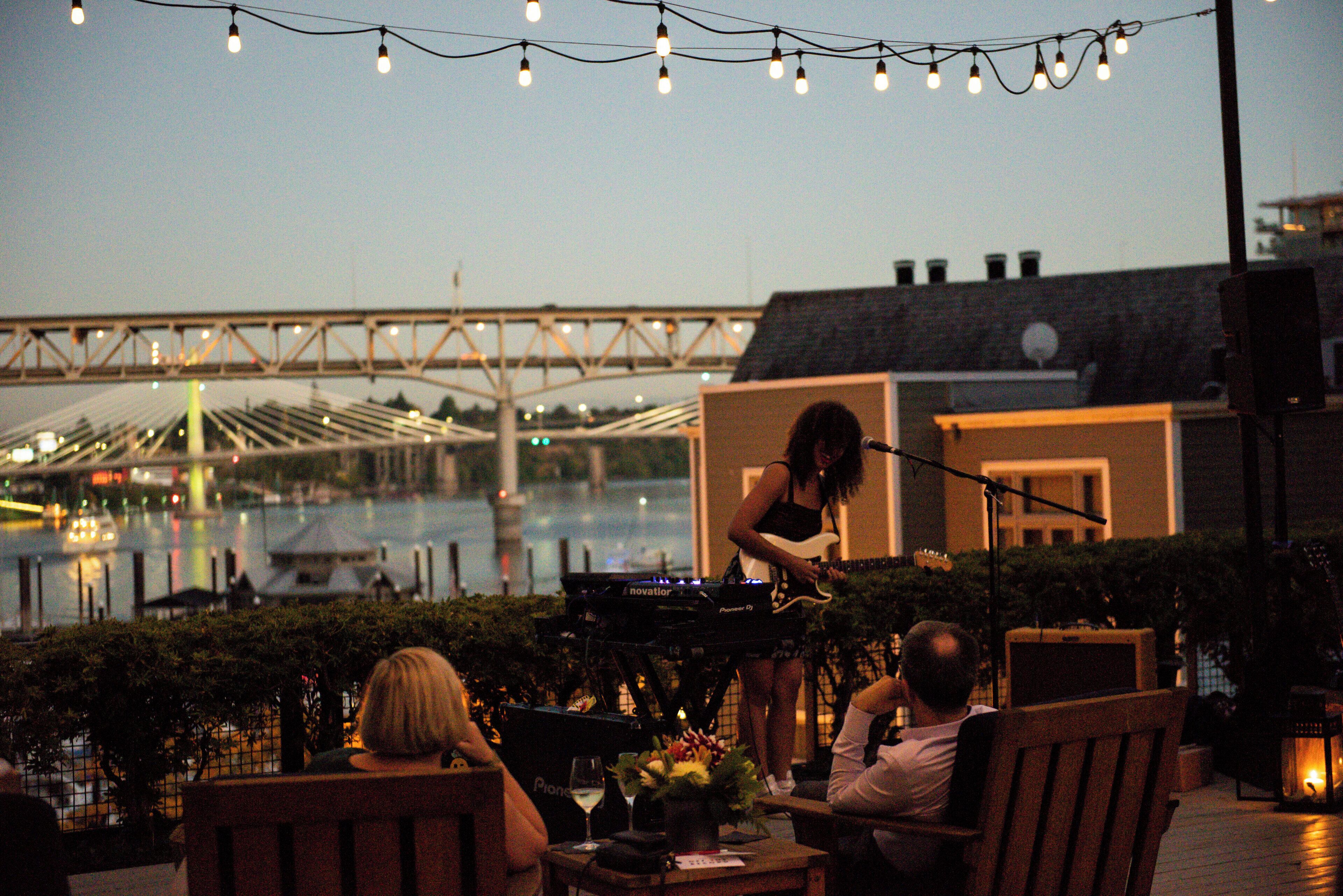 A musician performing on a keyboard at an outdoor venue with a view of a bridge and water in the background.