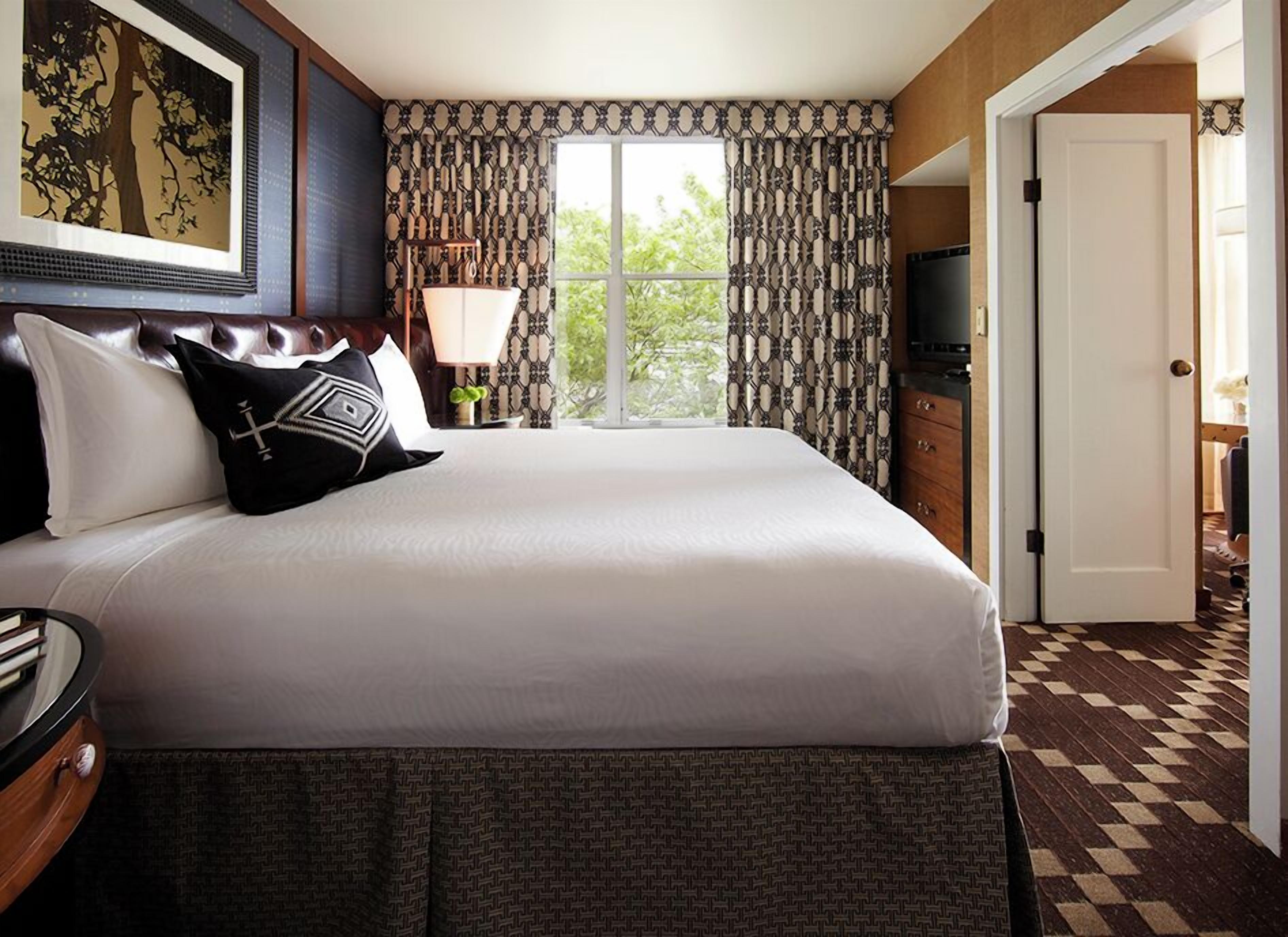A hotel room with a large bed, a dresser, a television, and a window with patterned curtains.