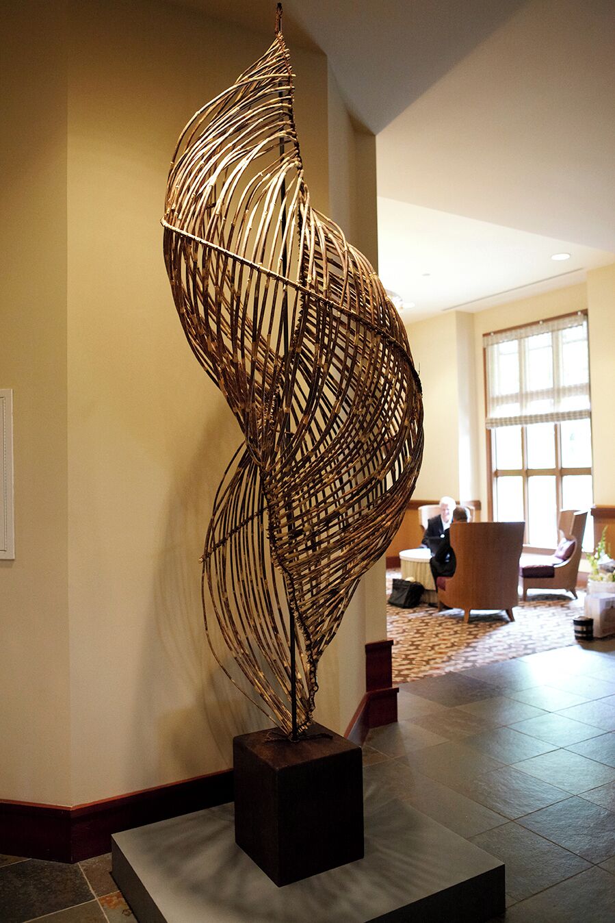Um lobby com uma grande escultura abstrata em um pedestal, uma área de estar com cadeiras, e uma janela ampla que oferece iluminação natural.