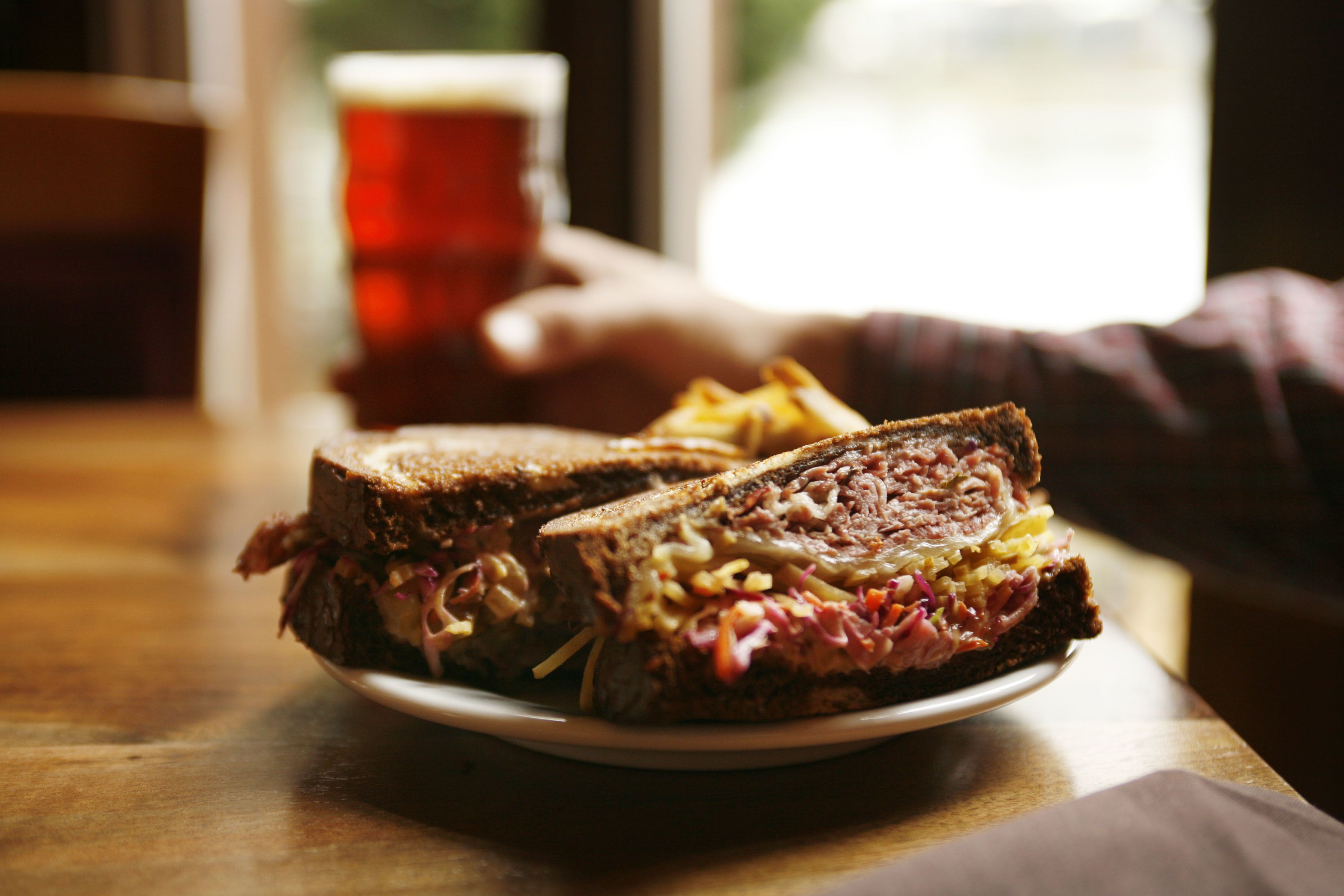 A sandwich on a plate with fries, a glass of beer, and a hand reaching for it.