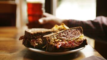 A sandwich on a plate with fries, a glass of beer, and a hand reaching for it.