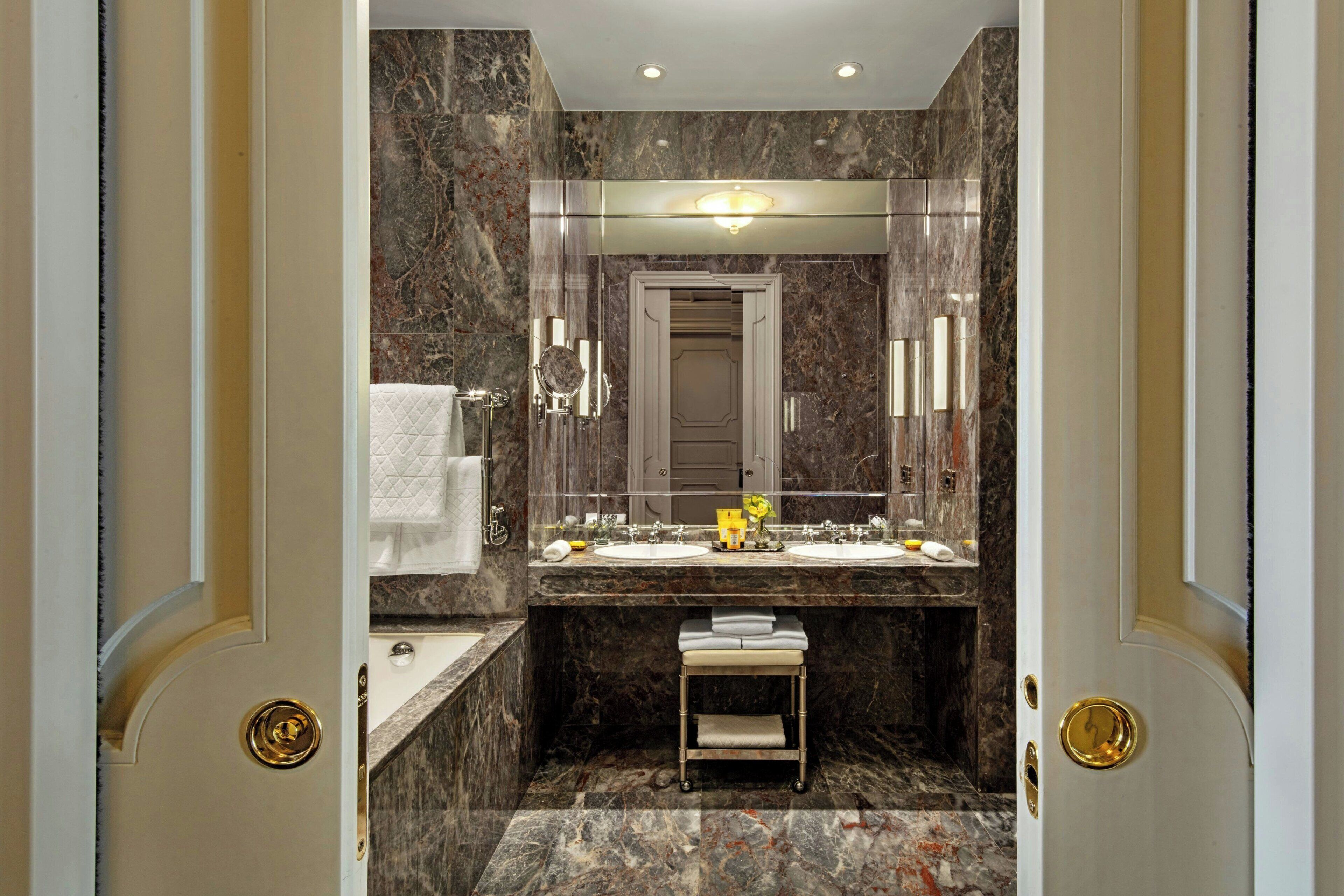 Un baño con paredes de mármol, un tocador con doble lavabo, un espejo y un taburete pequeño.