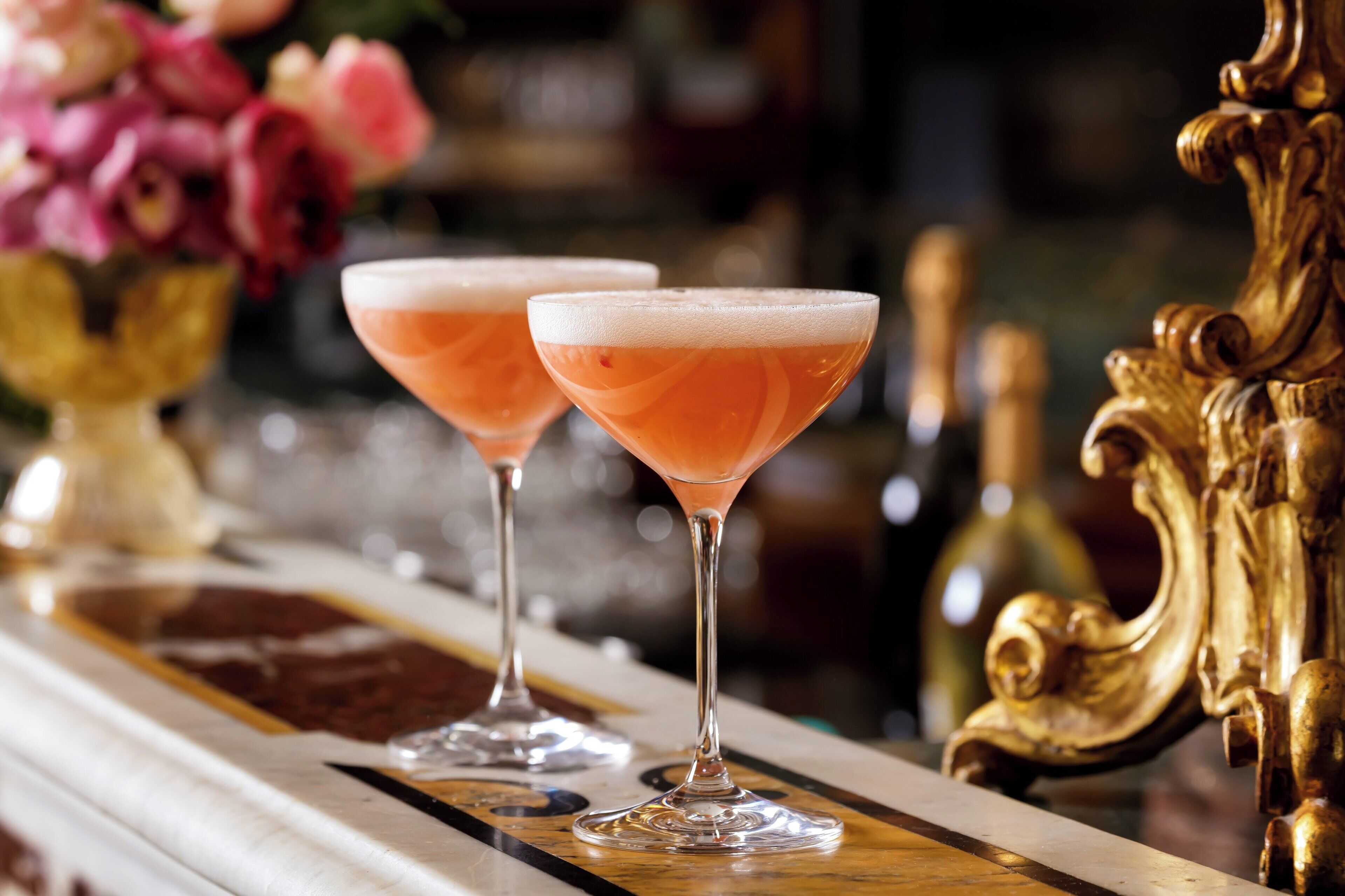 Two cocktails on a bar counter with a gold decorative element and a vase of flowers in the background.