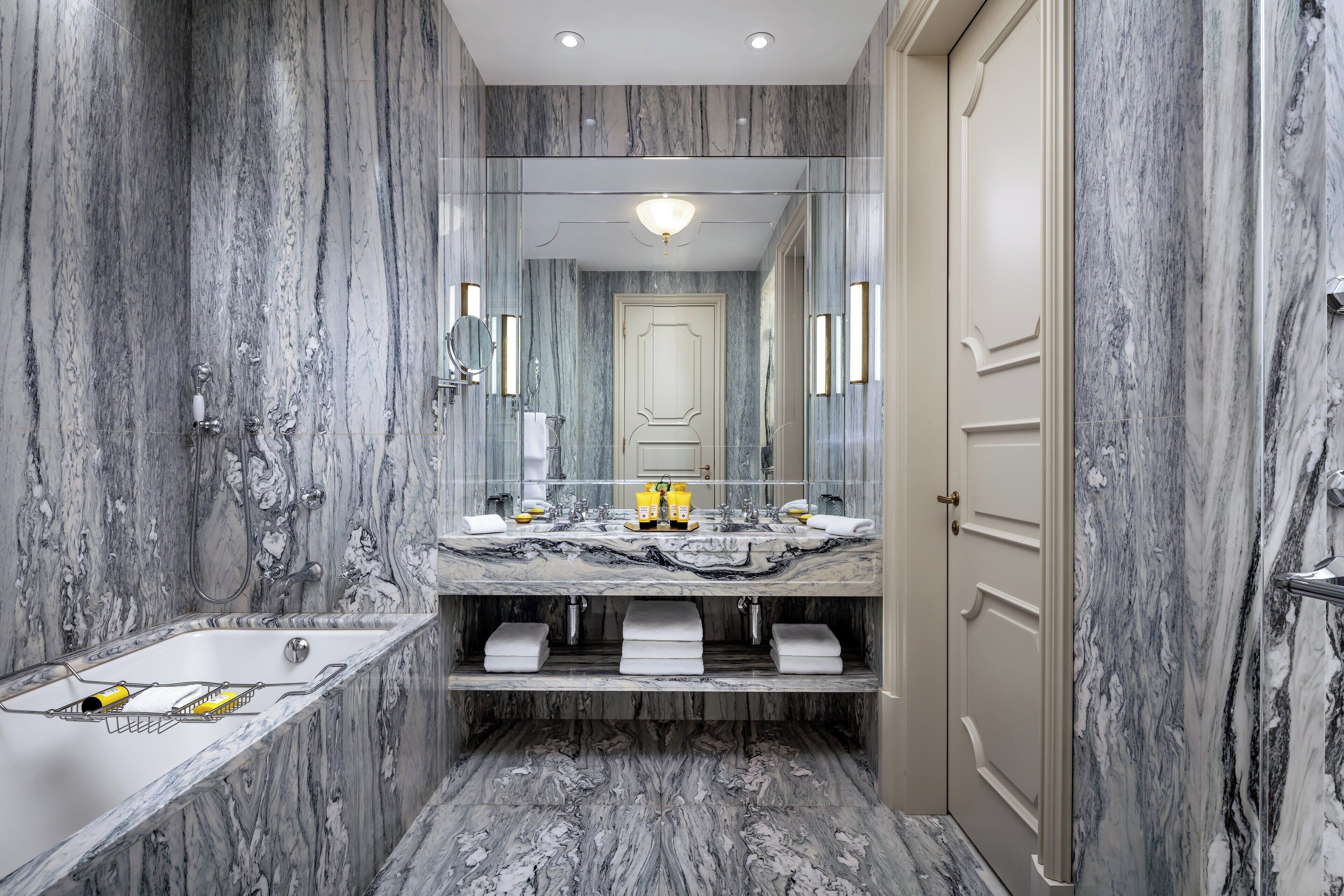 Une salle de bain avec des murs en marbre, un grand miroir, une baignoire et une porte.