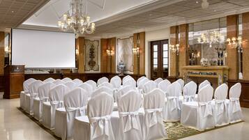 Un salón de conferencias con filas de sillas cubiertas de blanco, una pantalla grande y candelabros.