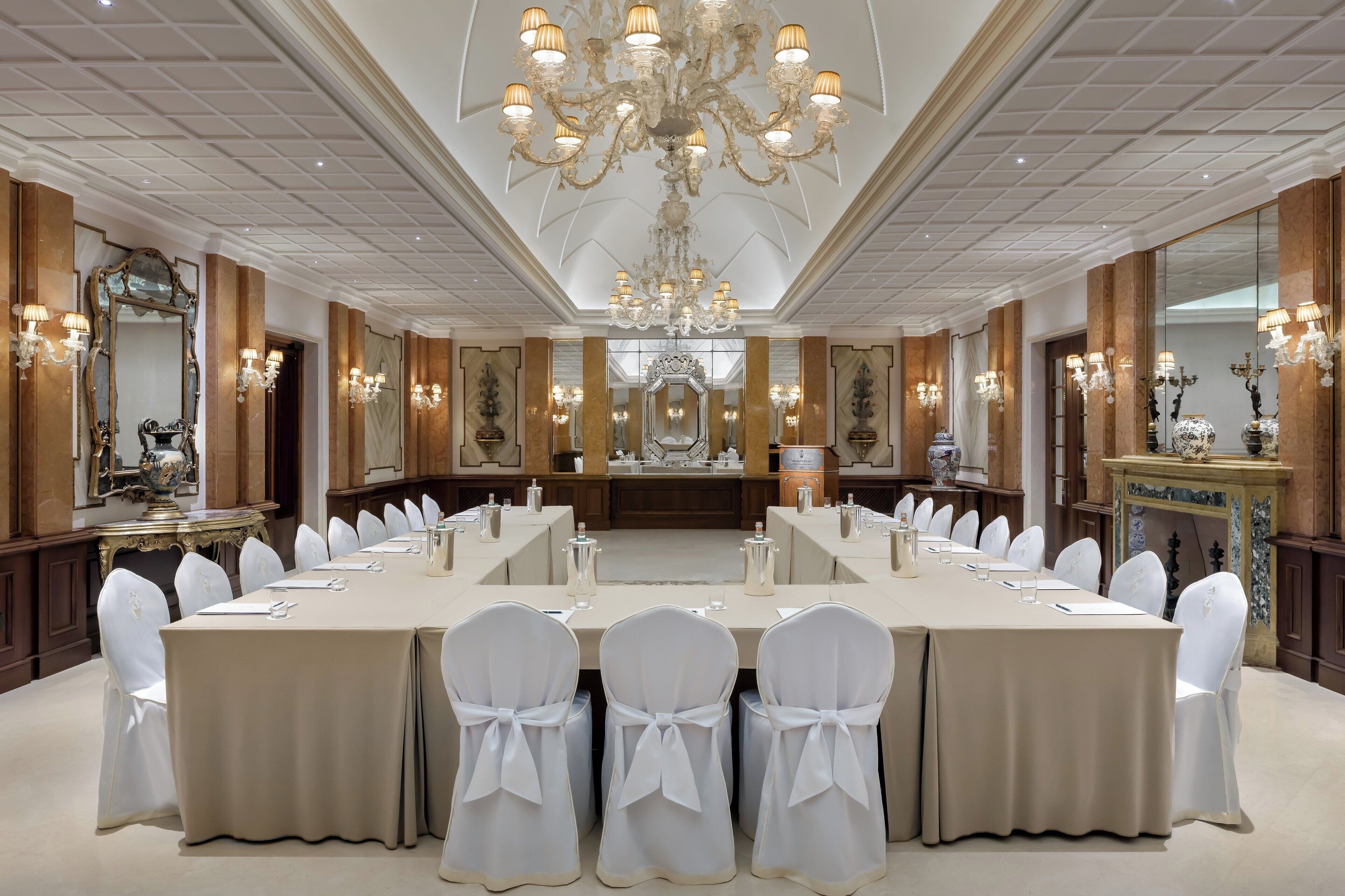 Un salón de banquetes elegante con una larga mesa dispuesta para una reunión, rodeado de sillas con cintas blancas y candelabros ornamentados.