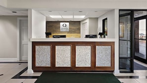 A hotel reception area with a modern design, featuring a reception desk, a sign that reads "Quality by Choice Hotels," and a view of the outside through a glass door.
