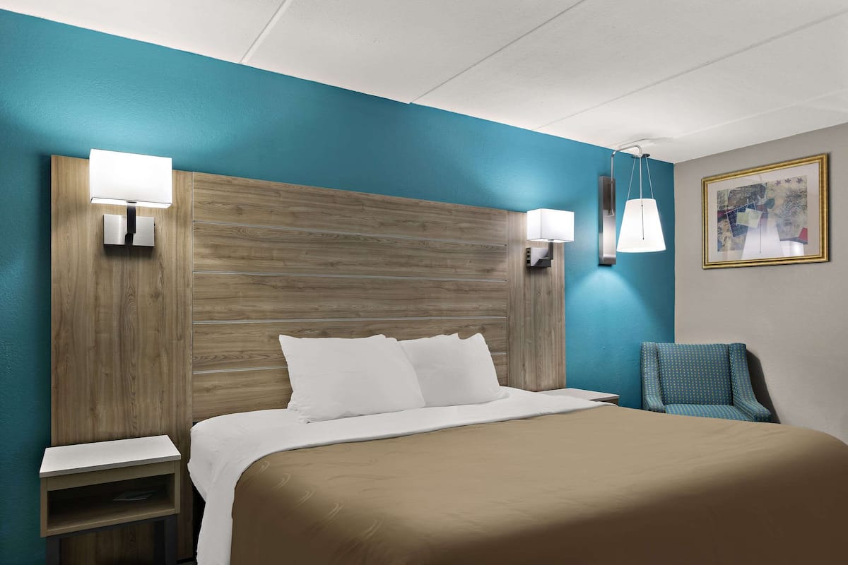 A hotel room with a wooden headboard, a bed with white linens, a nightstand, a blue armchair, and wall-mounted lamps.