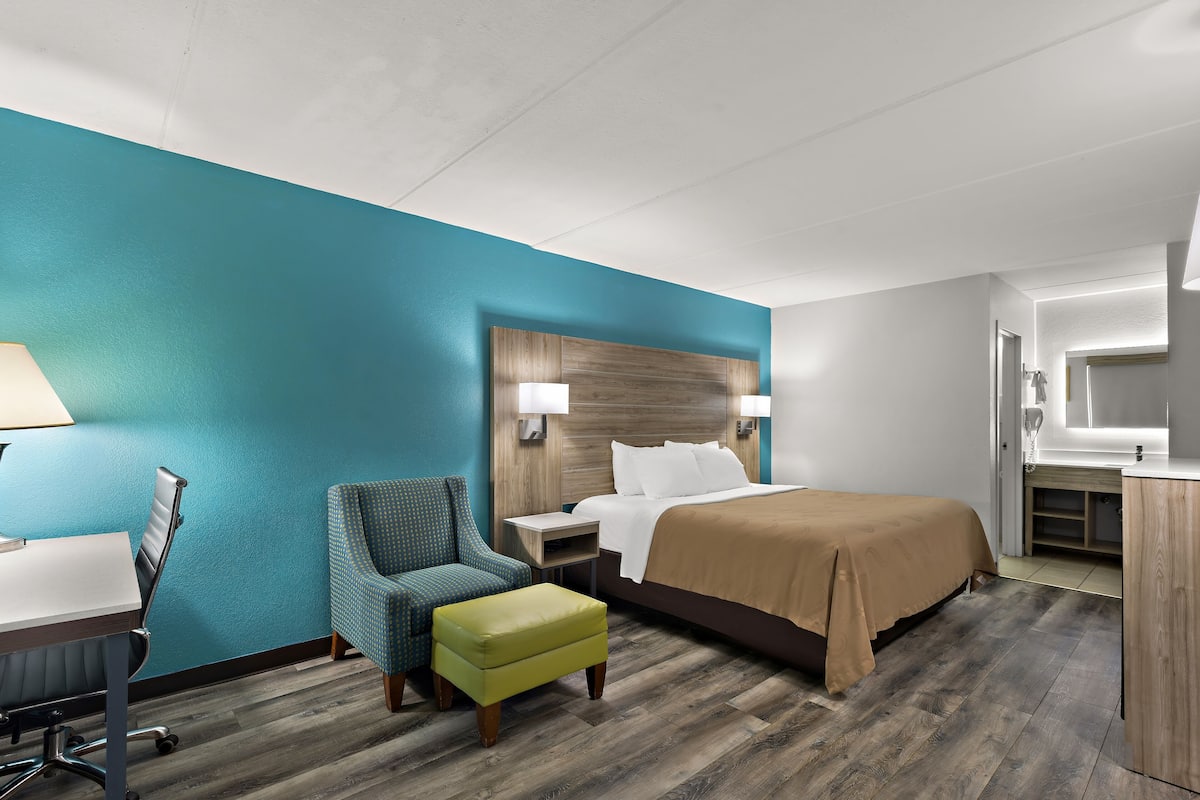 A hotel room with a teal accent wall, a wooden headboard, a bed with a beige blanket, a desk with a lamp, a chair, and a small ottoman.