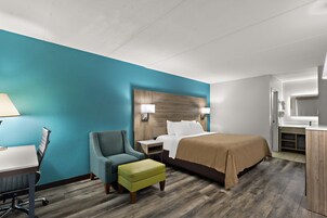 A hotel room with a teal accent wall, a wooden headboard, a bed with a beige blanket, a desk with a lamp, a chair, and a small ottoman.