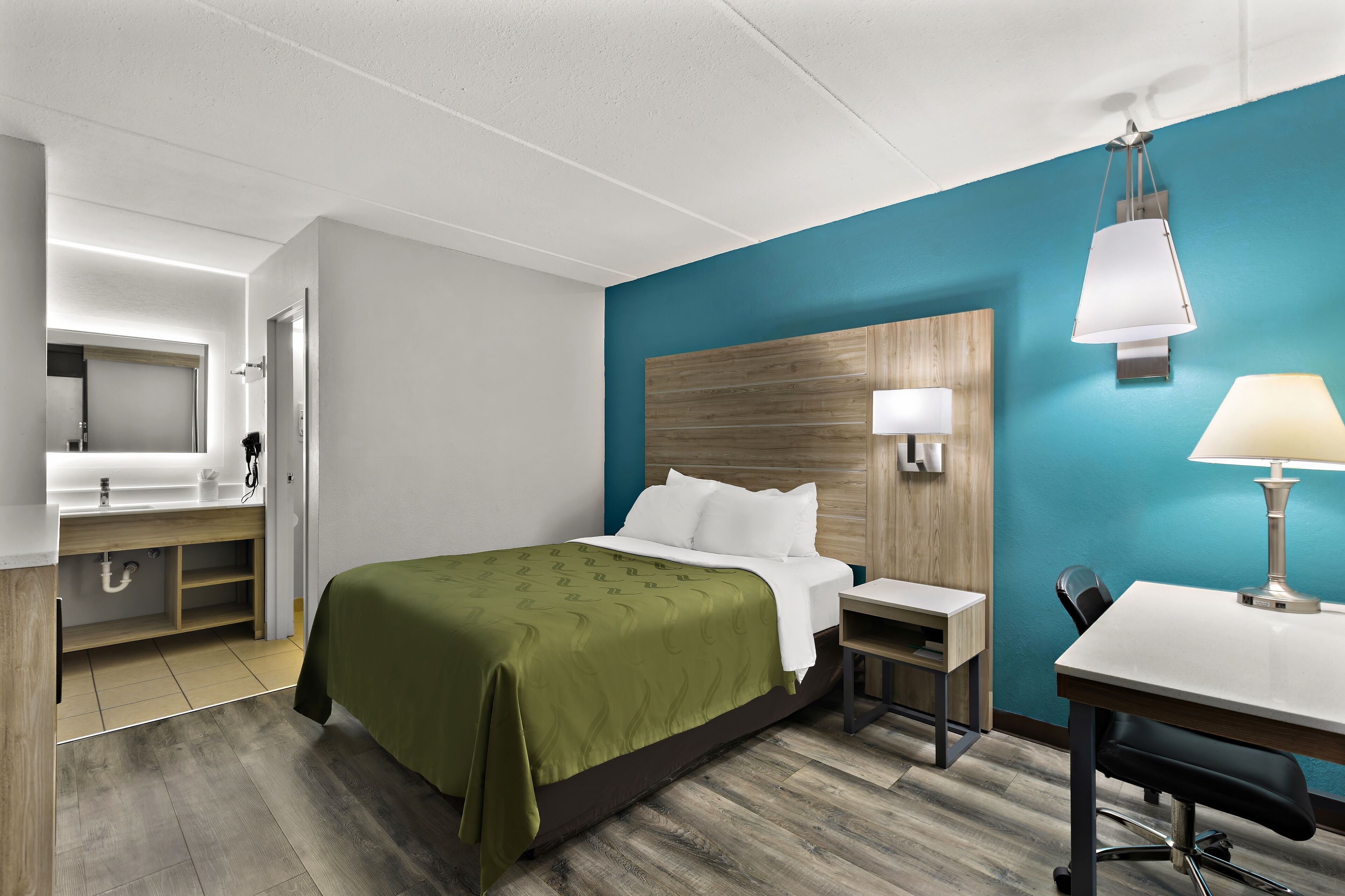A hotel room with a bed, a desk, a chair, a lamp, a mirror, and a wooden headboard.