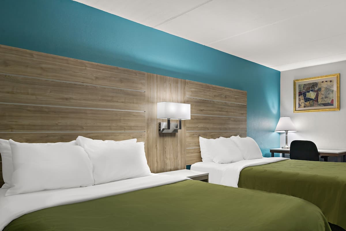 A hotel room with two beds, a wooden headboard, a desk with a chair, and a painting on the wall.
