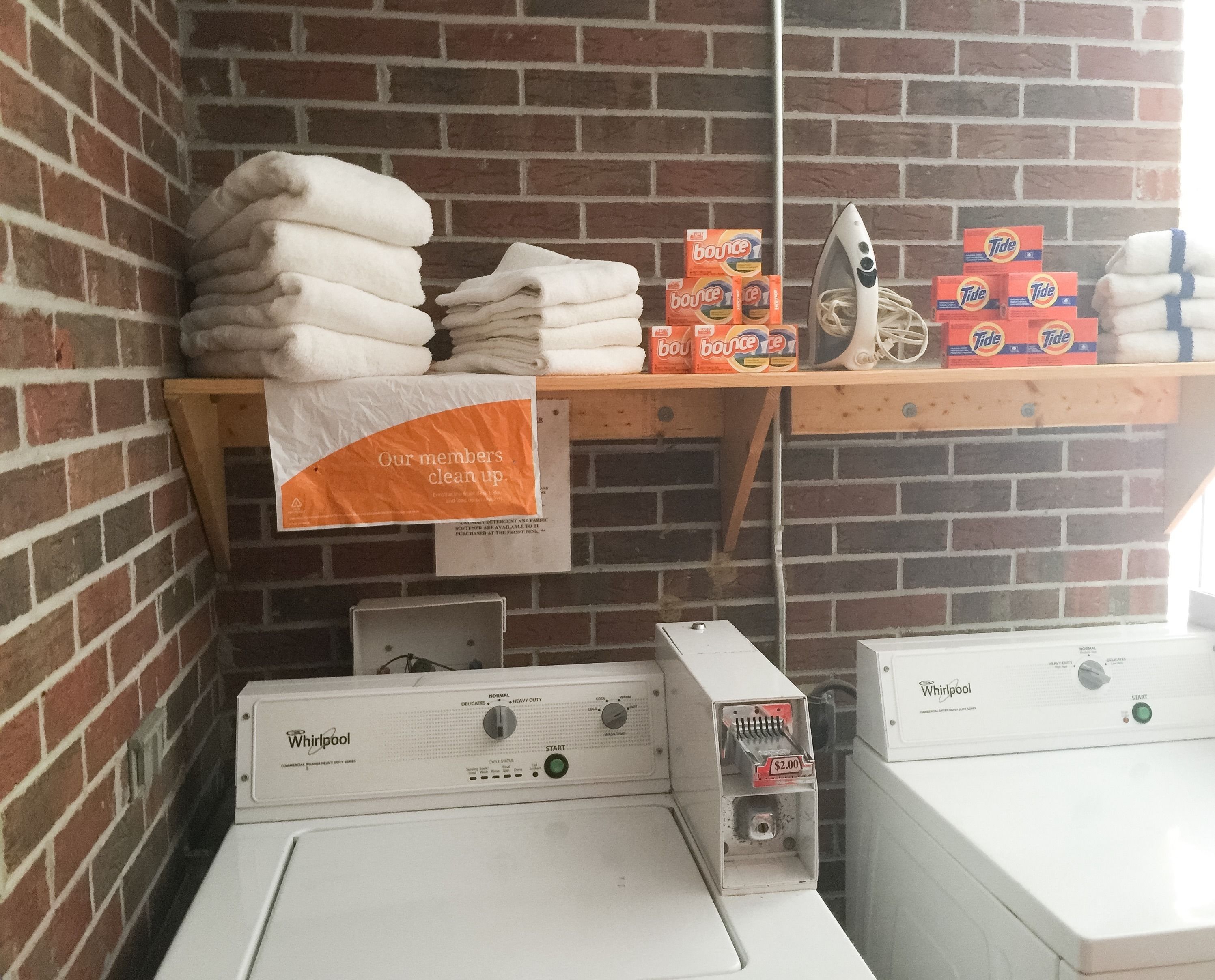 Zona de lavandería con toallas apiladas, cajas de detergente Tide y suavizante Bounce, un plancha y secadoras Whirlpool.