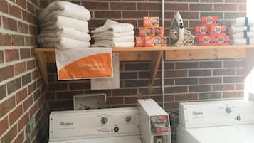 Zona de lavandería con toallas apiladas, cajas de detergente Tide y suavizante Bounce, un plancha y secadoras Whirlpool.