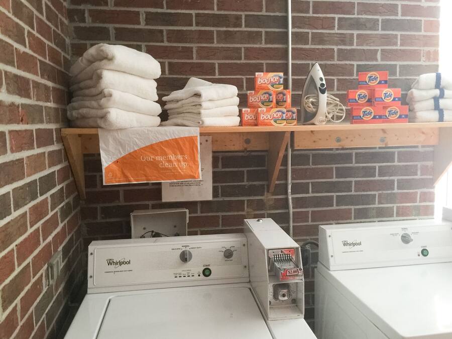Zona de lavandería con toallas apiladas, cajas de detergente Tide y suavizante Bounce, un plancha y secadoras Whirlpool.