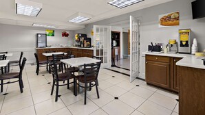 A clean, modern restaurant interior with white tables and chairs, a kitchen area, and a coffee station.