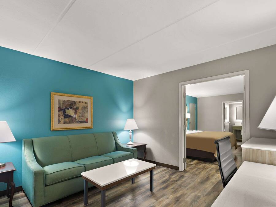 Una habitación de hotel con pared verde azulado, sofá verde, mesa blanca, cama con edredón color beige y un espejo.