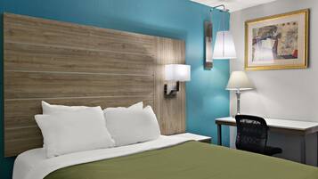 Una habitación de hotel con cabecera de madera, un escritorio con silla, una lámpara y un cuadro en la pared.
