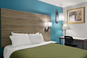 A hotel room with a wooden headboard, a desk with a chair, a lamp, and a painting on the wall.