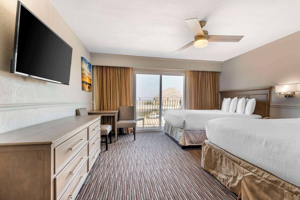 A hotel room with a flat-screen TV, a desk, two beds, a ceiling fan, and a view of the outdoors.