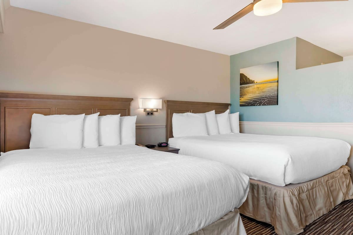 A hotel room with two beds, a ceiling fan, a nightstand with a lamp, and a painting on the wall.