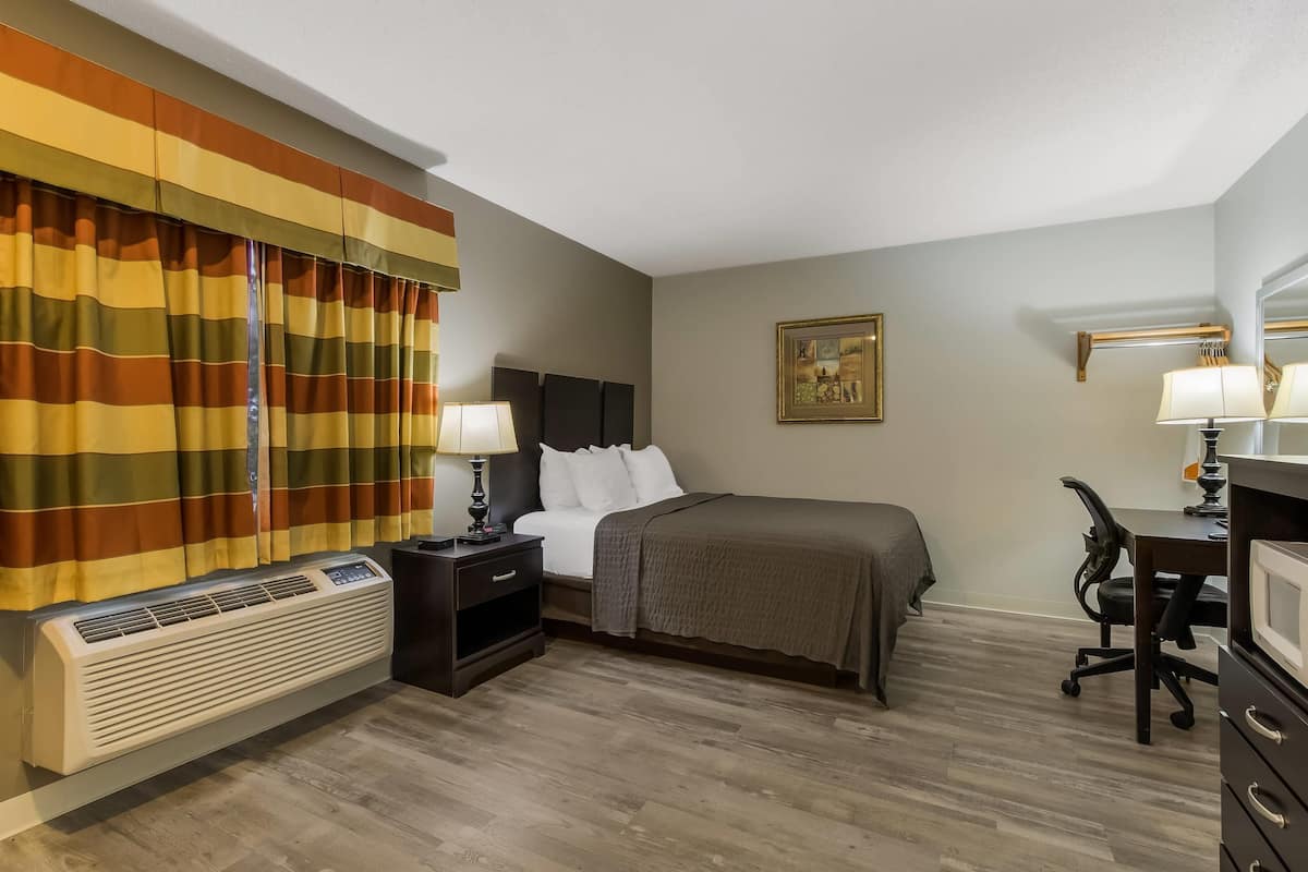 A hotel room with a bed, bedside tables, a desk, a chair, a heater, and a window with curtains.