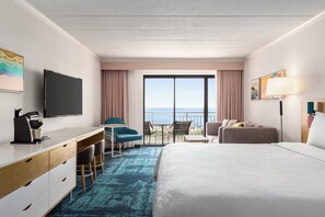 A hotel room with a large bed, a desk with a chair, a TV, and a balcony with a view of the sea.