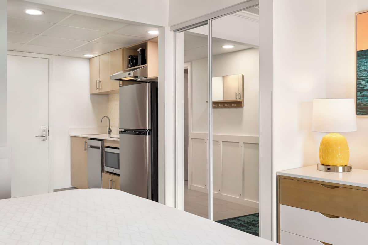 A hotel room with a bed, a desk, a lamp, a bathroom, and a kitchenette.