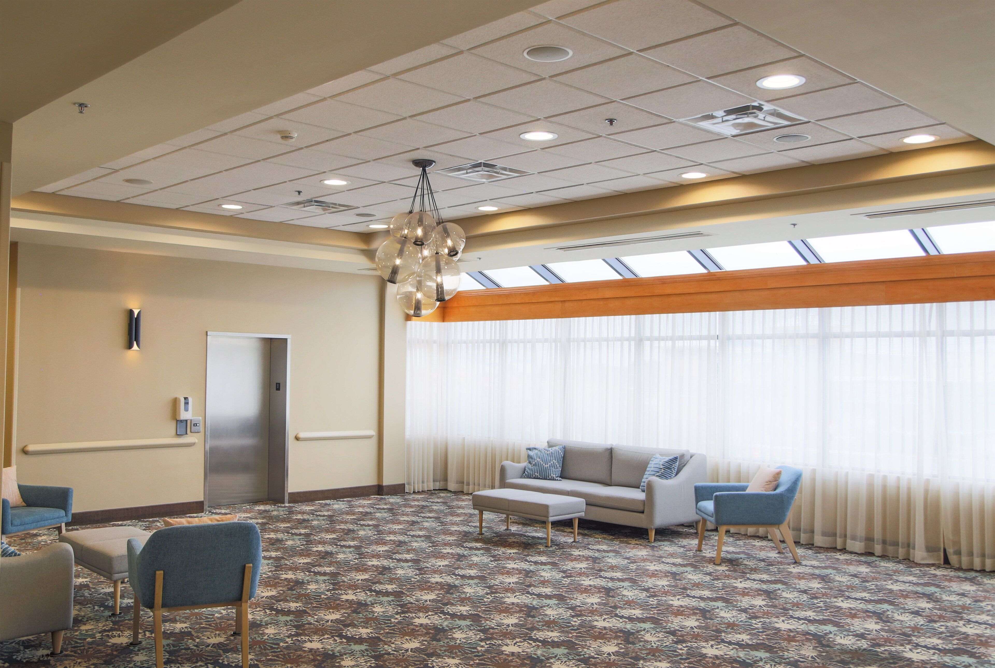 A hotel lobby with a patterned carpet, a sofa, two armchairs, a coffee table, a chandelier, a door, and large windows with curtains.