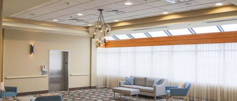 A hotel lobby with a patterned carpet, a sofa, two armchairs, a coffee table, a chandelier, a door, and large windows with curtains.