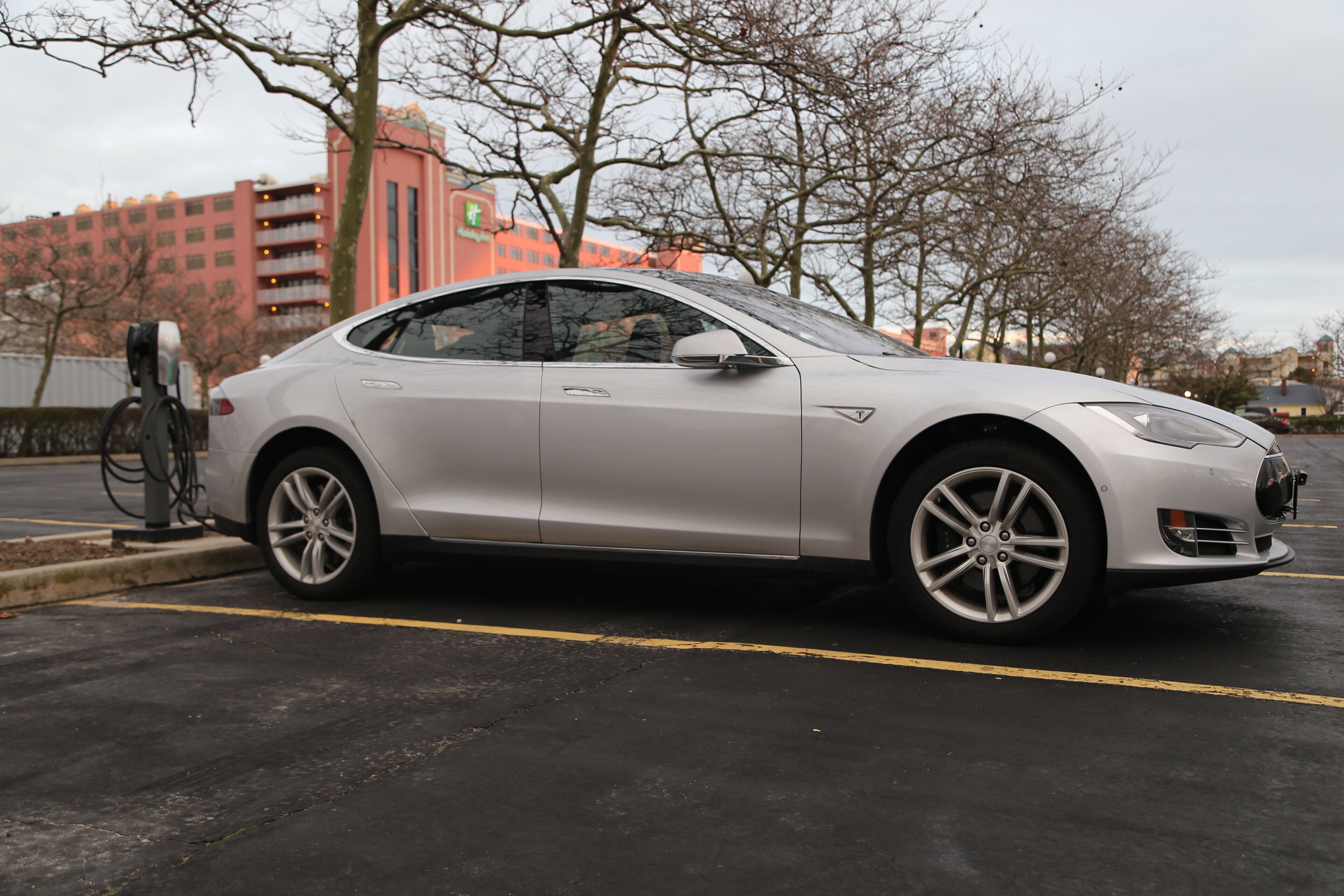 A silver Tesla parked in a lot with a charging station, trees, and buildings in the background.