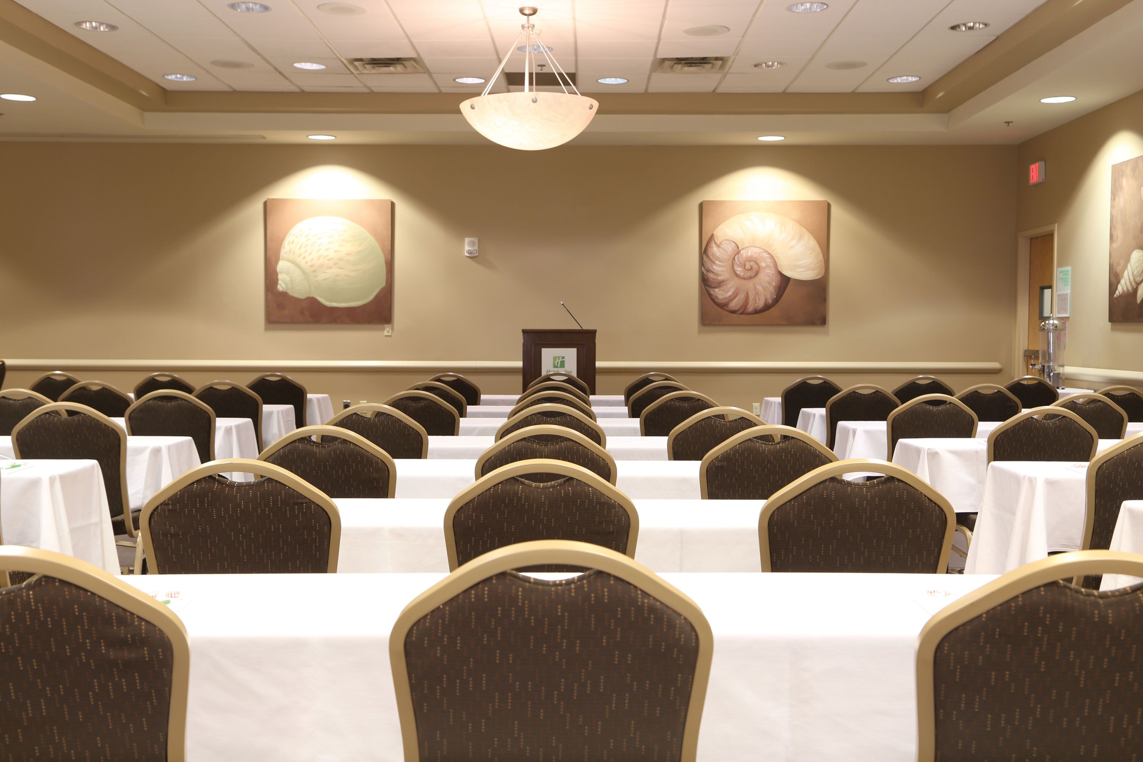 A conference room with rows of tables and chairs, a podium, and two large shell paintings on the walls.