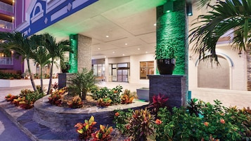 Entrada de hotel com fachada de pedra, parede iluminada em verde e diversas plantas tropicais.