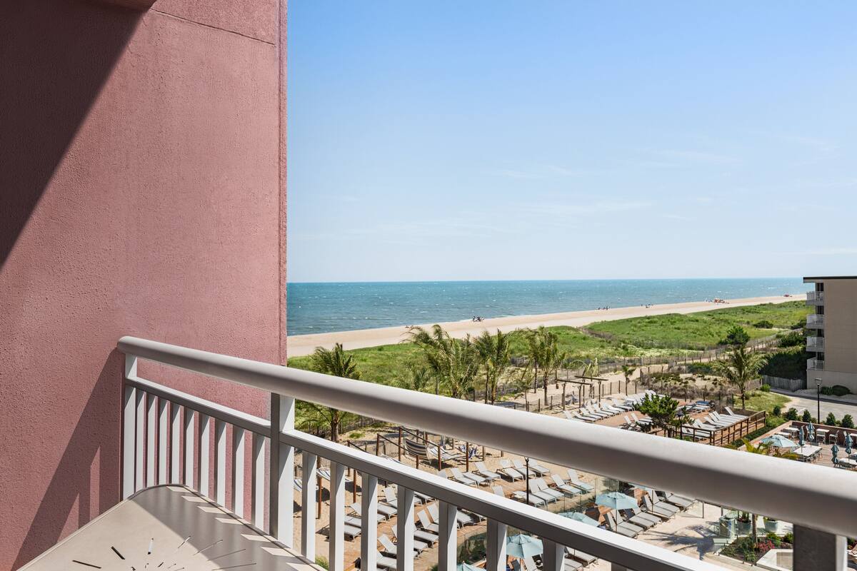 A balcony with a view of a beach, lounge chairs, and a fence separating the beach from the resort area.