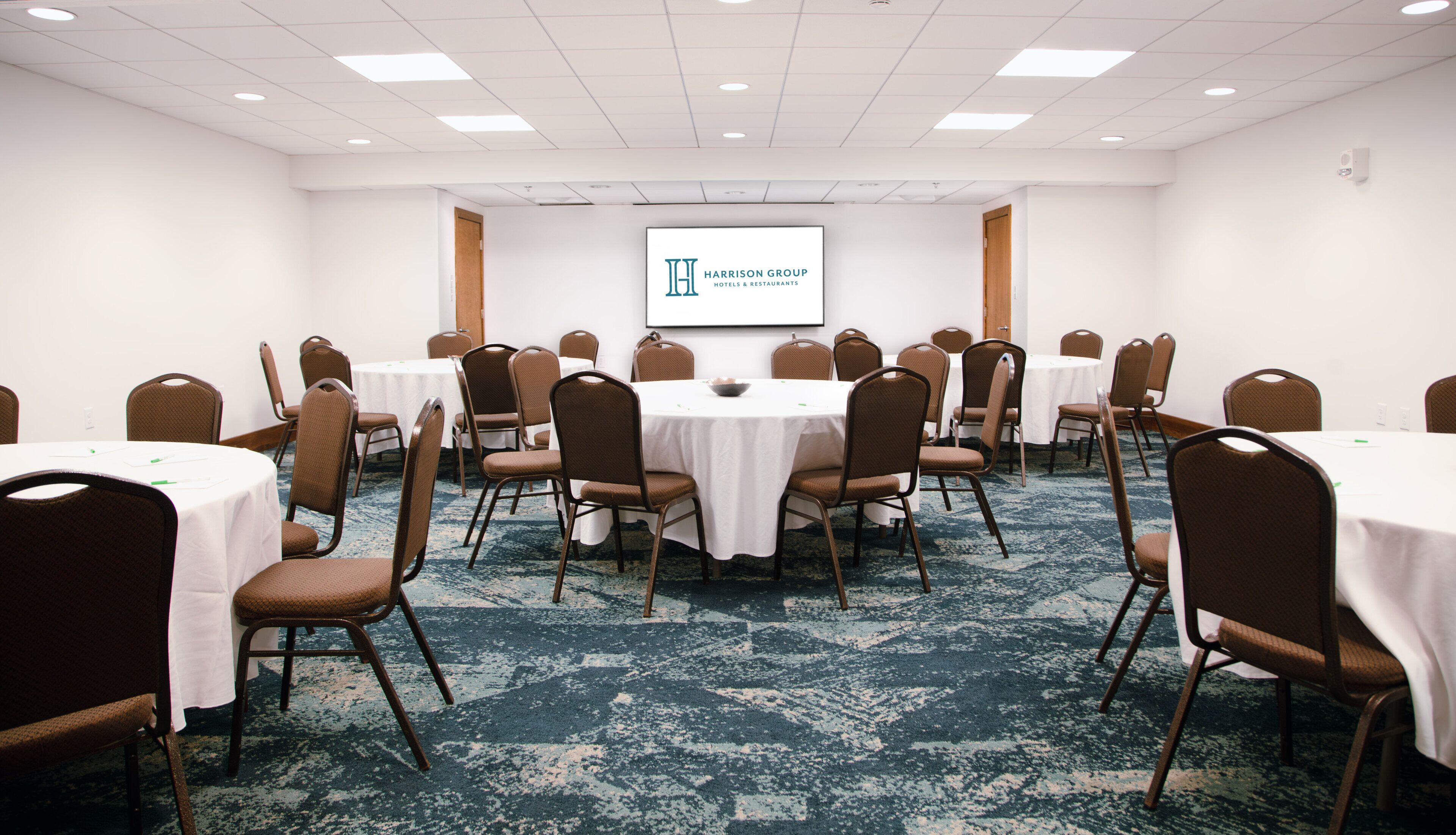 Un salón de conferencias con mesas redondas, sillas marrones y una pantalla de proyector que muestra 'Harrison Group Hotels & Restaurants'.