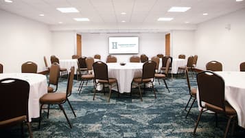 Un salón de conferencias con mesas redondas, sillas marrones y una pantalla de proyector que muestra 'Harrison Group Hotels & Restaurants'.