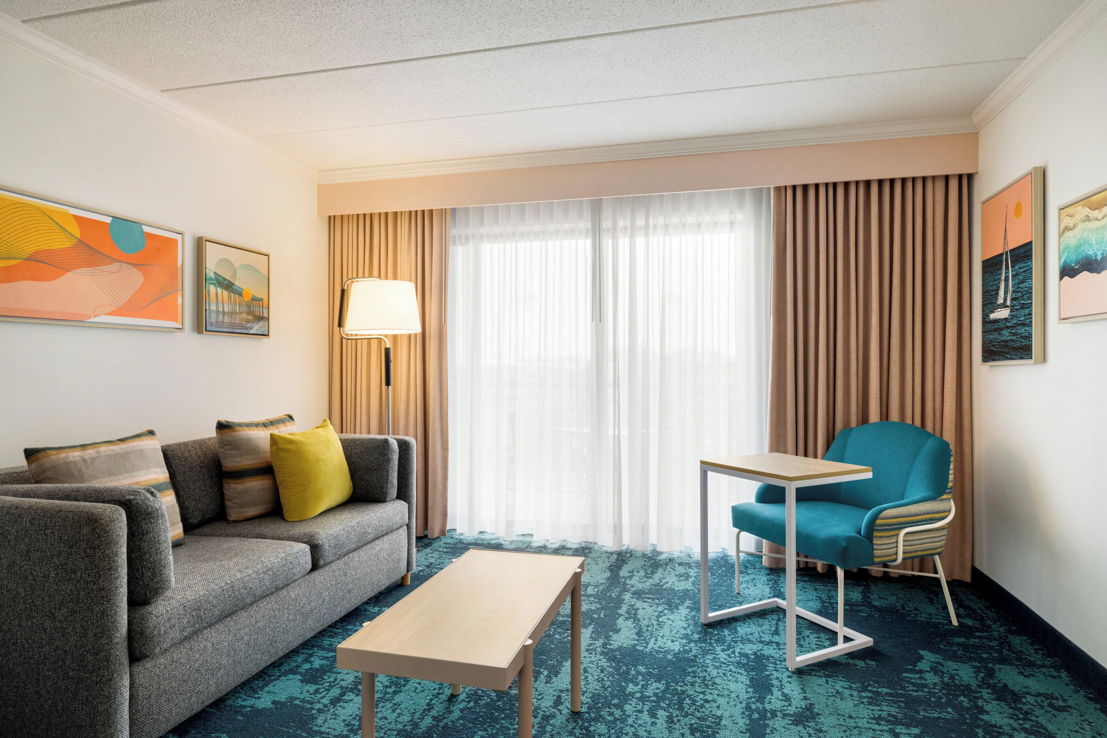 Habitación de hotel con un sofá gris, una mesita pequeña, un sillón azul aguamarina y un ventanal con cortinas.