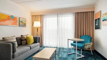 Habitación de hotel con un sofá gris, una mesita pequeña, un sillón azul aguamarina y un ventanal con cortinas.