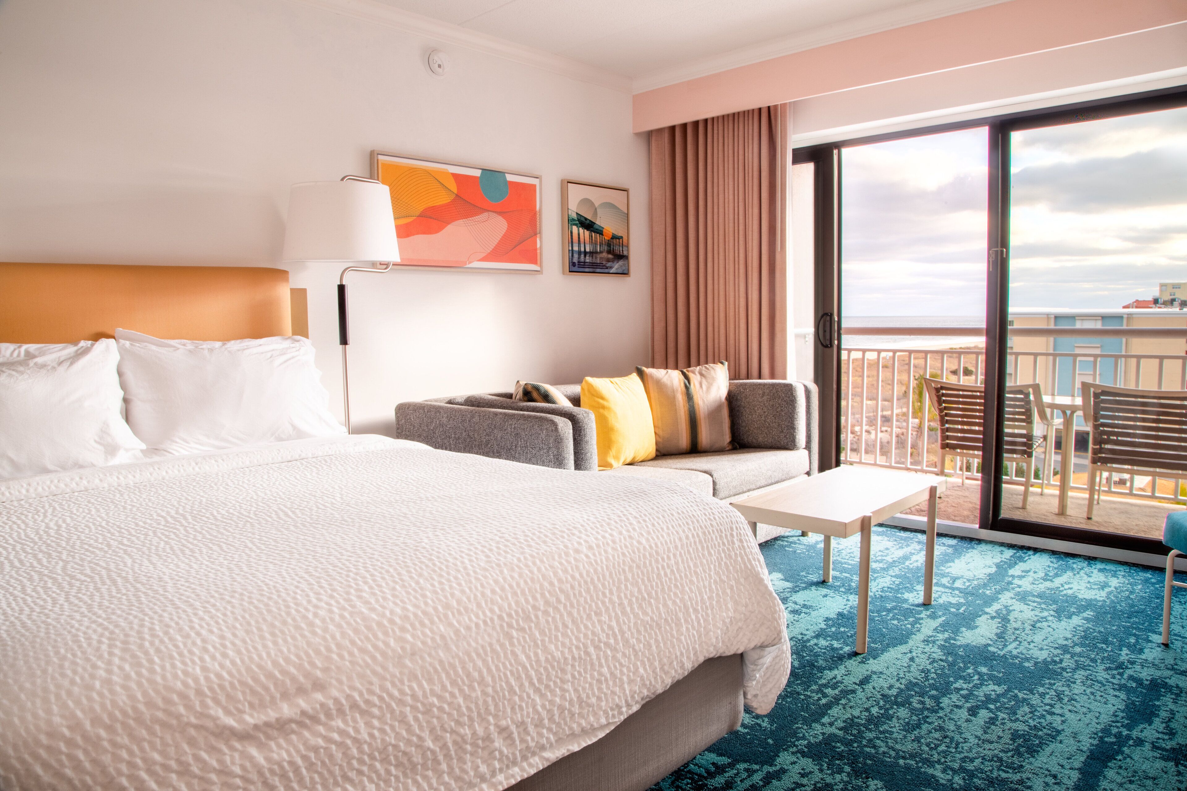 A hotel room with a large bed, a sofa, a small table, and a balcony with a view of the beach.