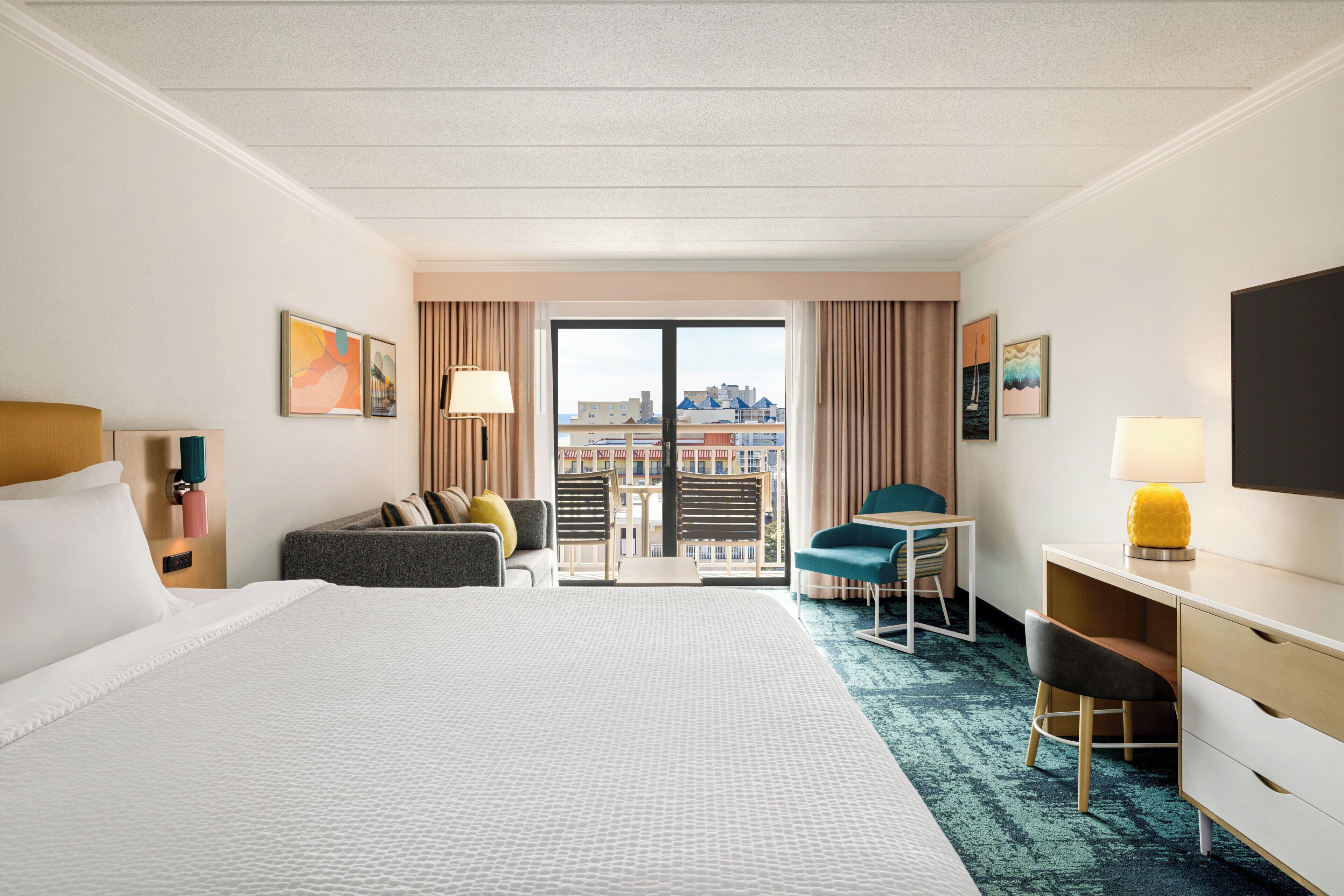 A hotel room with a large bed, a desk, a chair, a small table, and a view of the city through a glass door.
