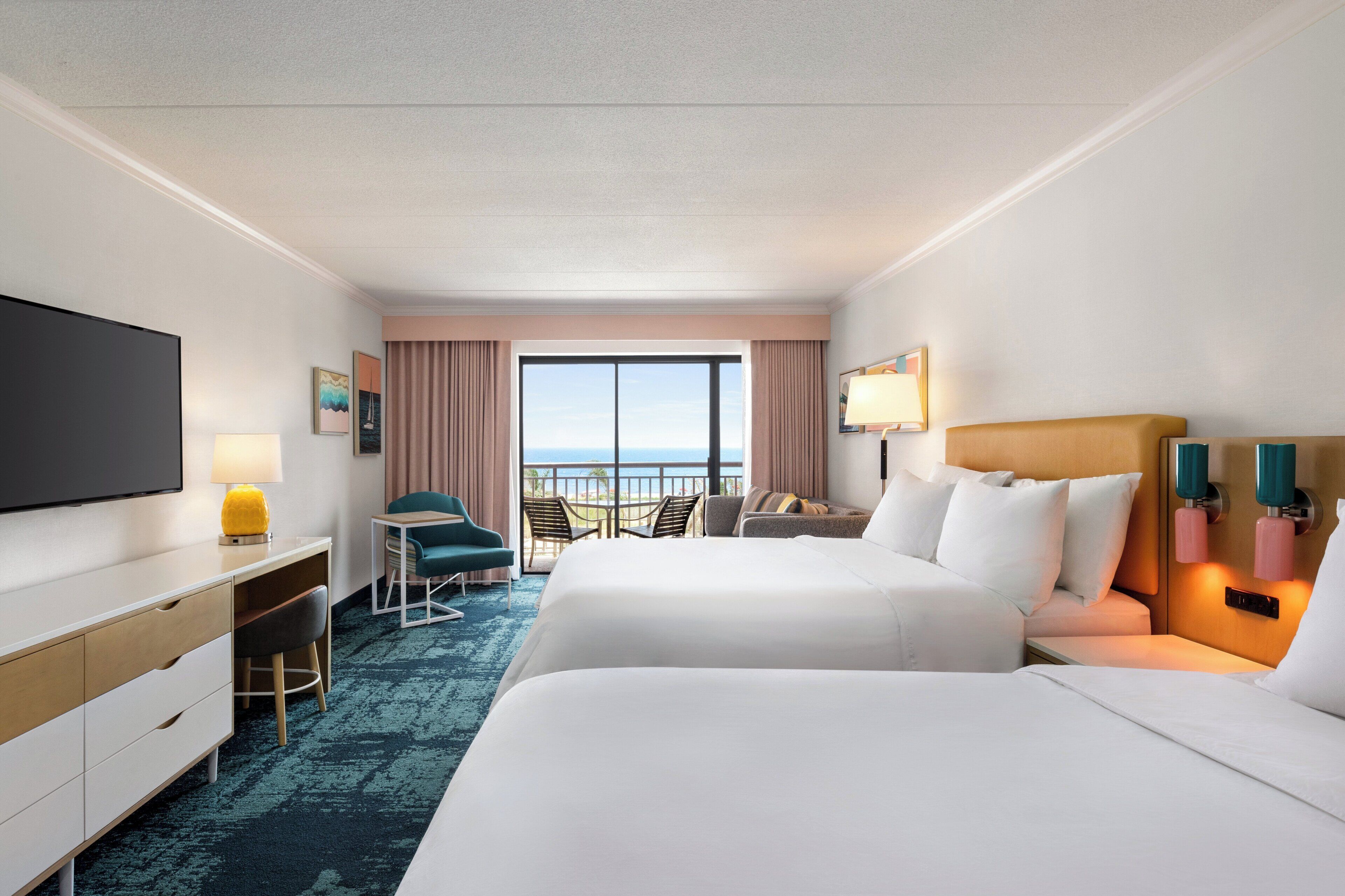 A hotel room with two beds, a desk, a chair, a TV, and a balcony with a view.