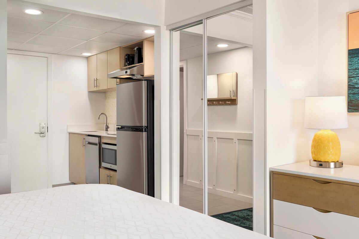 A hotel room with a bed, a desk, a lamp, a bathroom, and a kitchenette.