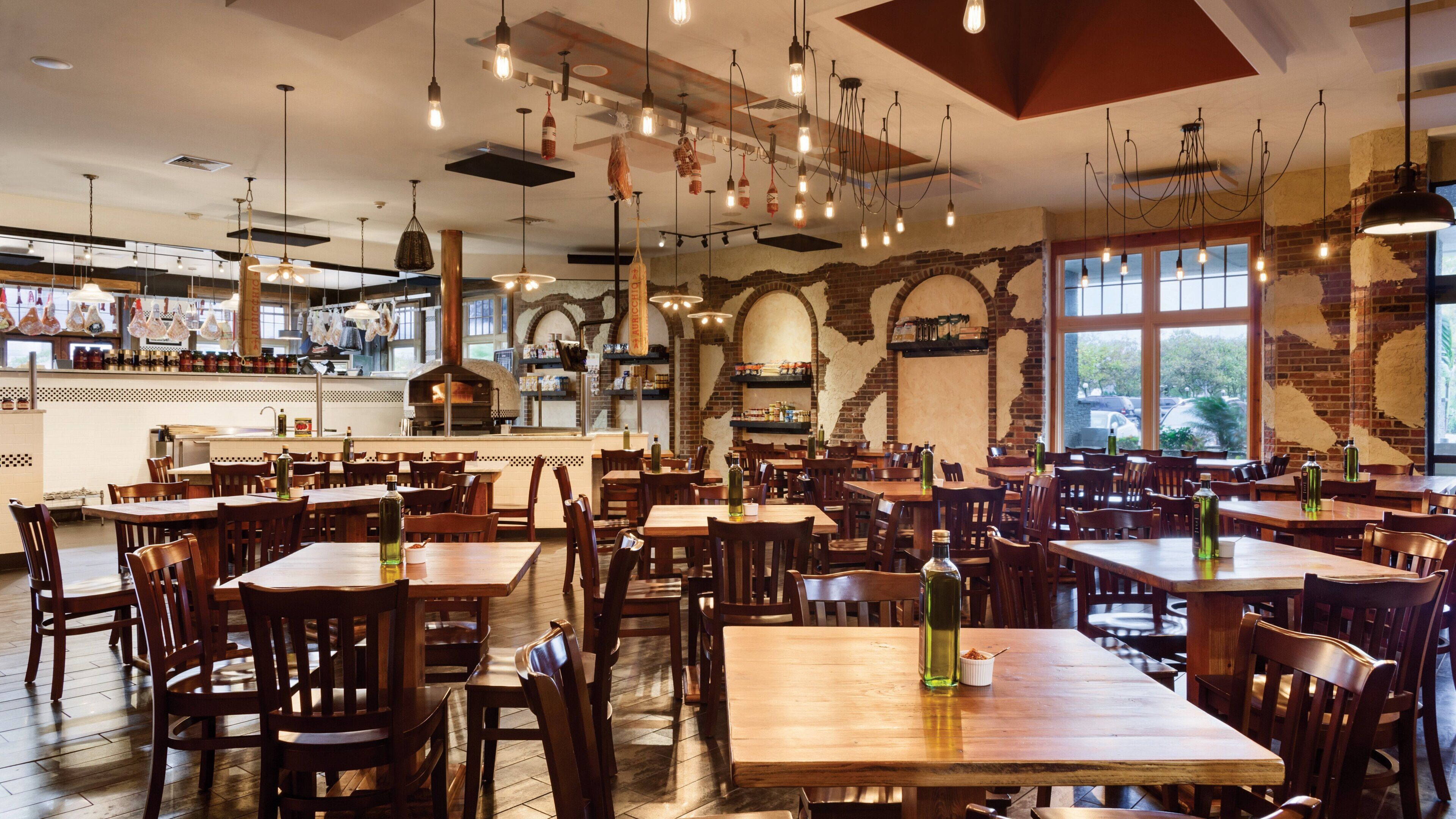 A restaurant with multiple wooden tables and chairs, hanging light fixtures, and a brick wall with a window.