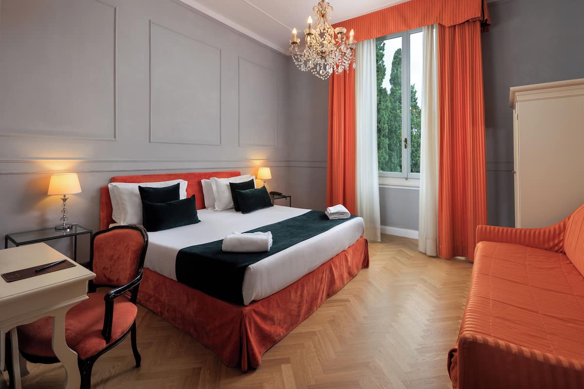 A hotel room with a large bed, two bedside tables with lamps, a desk, a chair, and a view of the outdoors through a window with orange curtains.
