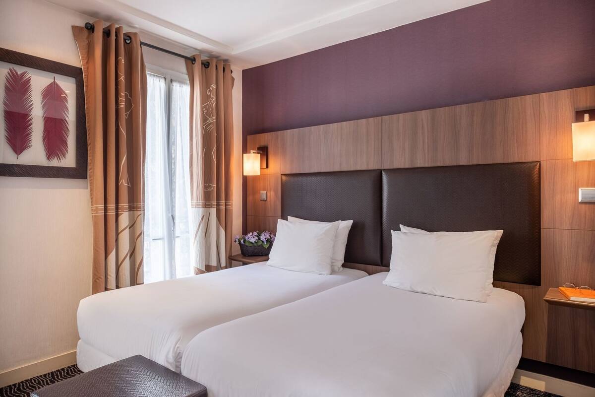 A hotel room with two beds, a nightstand with a lamp, a window with curtains, and a wall-mounted artwork.