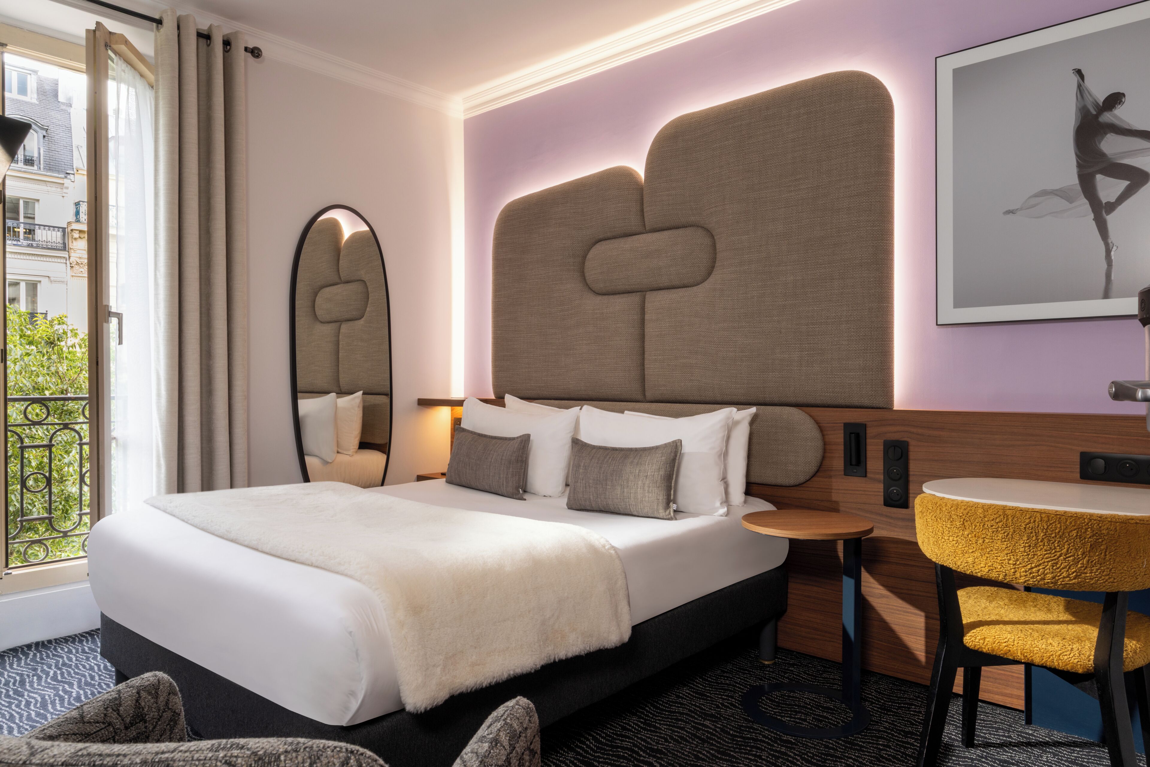 A hotel room with a large bed, a round table, a chair, a mirror, and a city view.