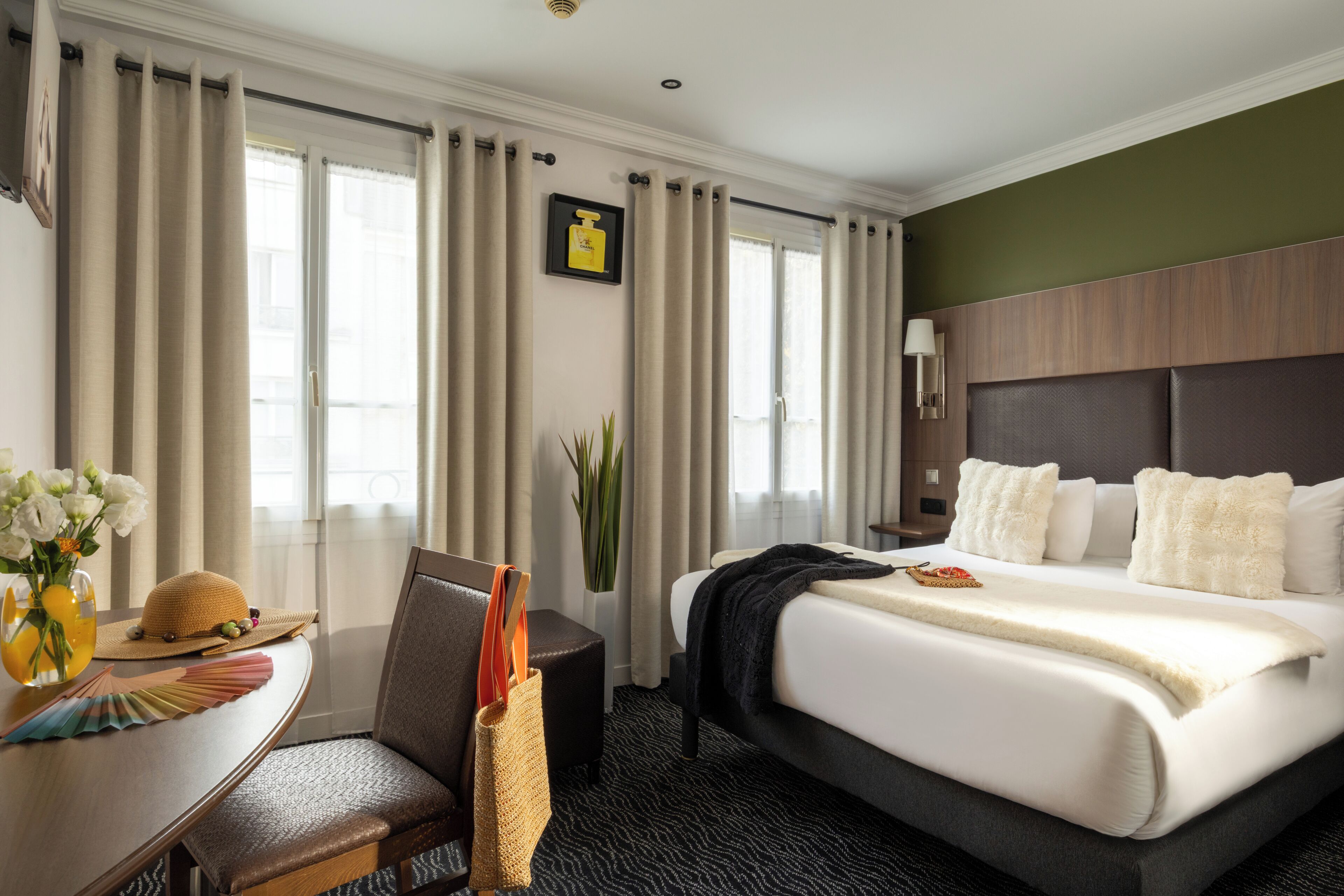 A hotel room with a bed, a desk with a chair, a vase with flowers, and a plant.