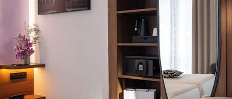 A hotel room with a bed, a bedside table with a lamp, a shelf with a coffee maker, a mirror reflecting the room, and a window with curtains.