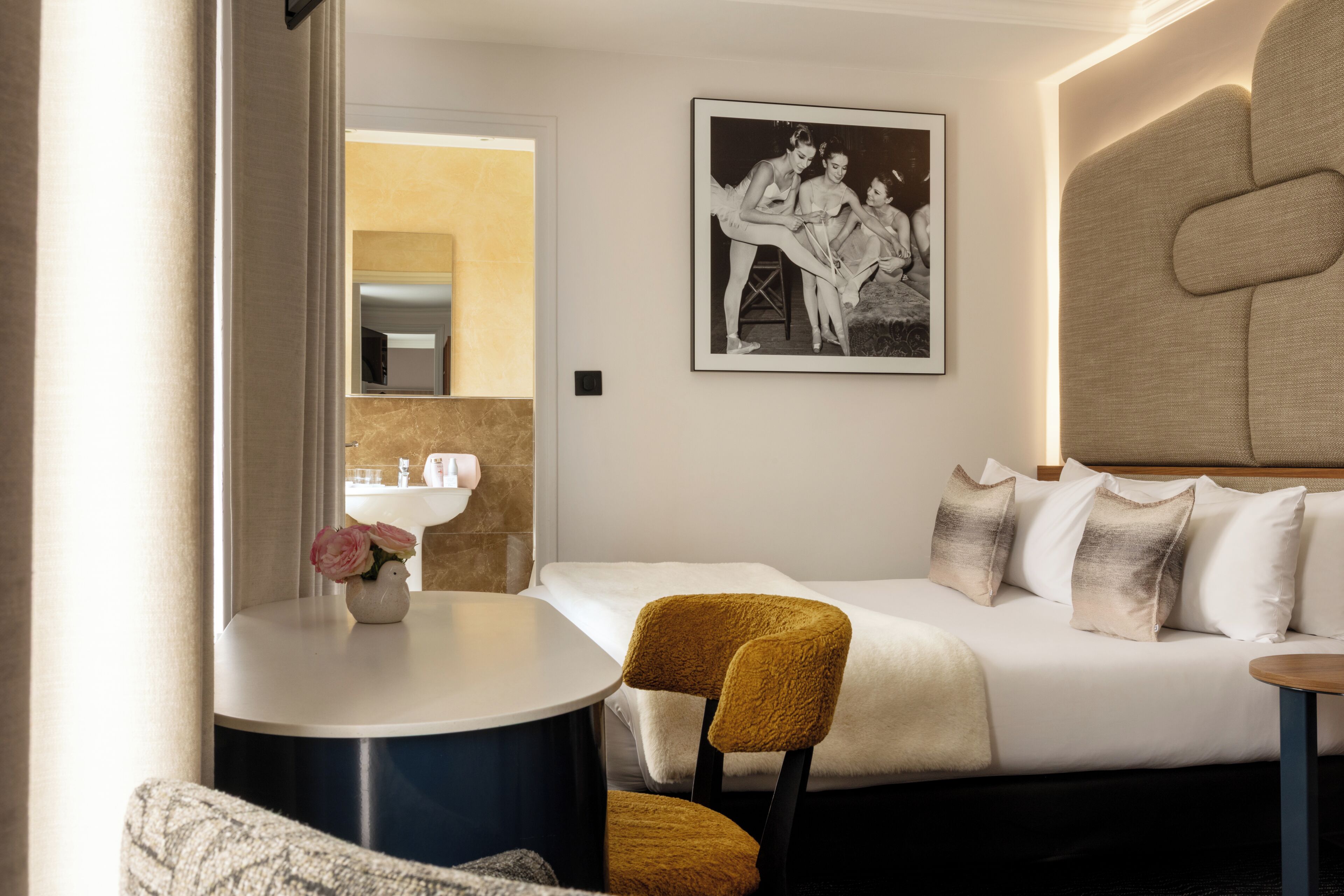 A hotel room with a bed, a sofa, a round table, and a chair. There is a framed picture on the wall and a vase with flowers on the table.